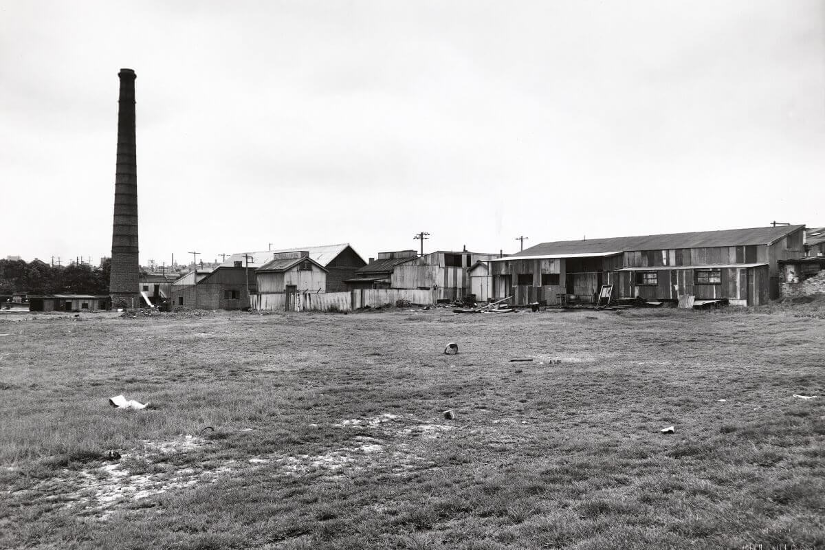 Black and white photo of Camdenville Park, St Peters, taken in the 1950s. The view looks northeast across a soil holding depot near Council Street, with a chimney stack from a nearby brickworks visible in the background. Several houses line the back of the photo. The image shows the site before the construction of the sports area where the first Koori Knockout would later take place