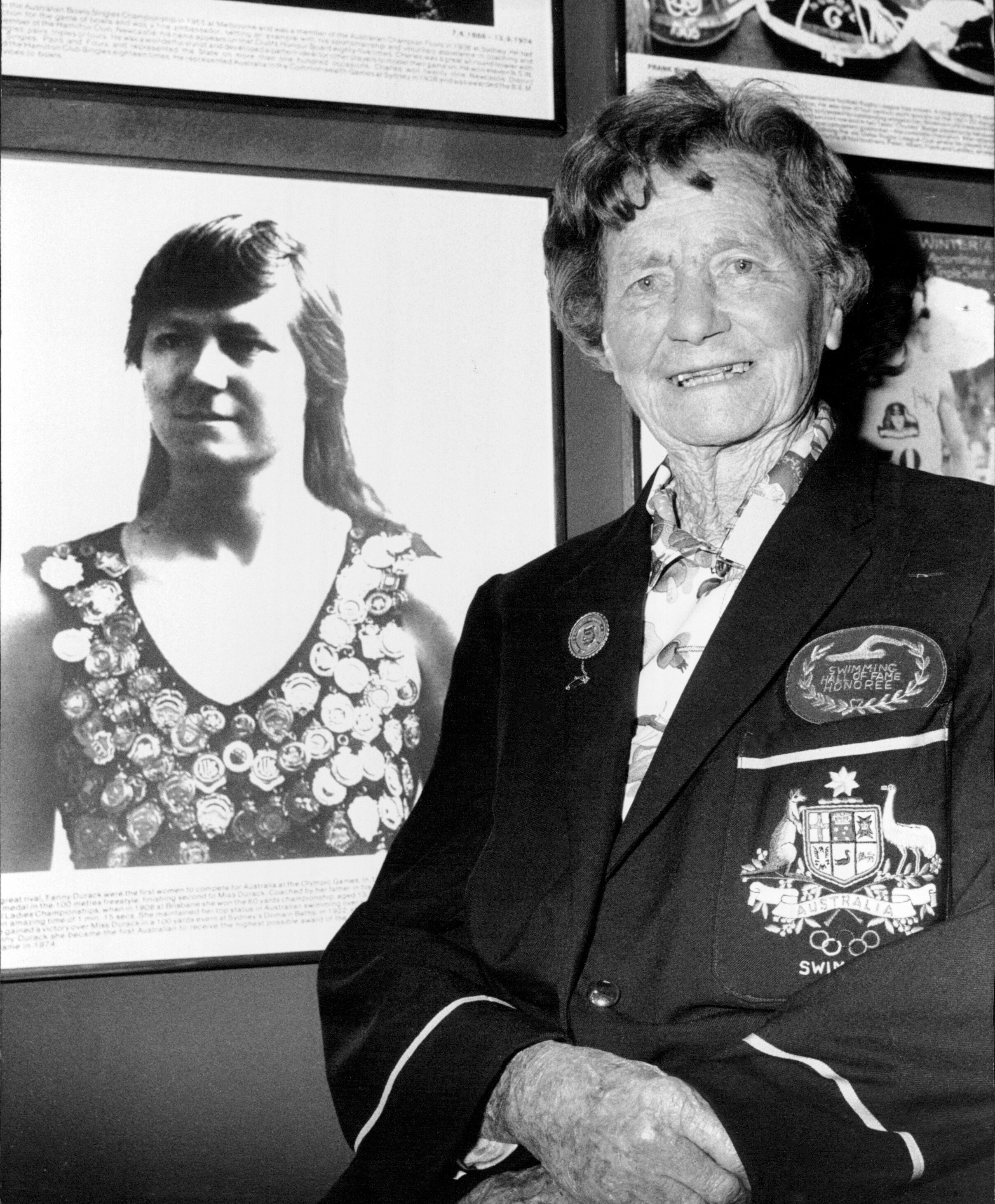 An elderly woman stands beside a portrait of her younger self in the Hall of Champions. The portrait shows the younger woman wearing a vest decorated with medals or badges. The elderly woman wears a blazer with an embroidered emblem and several pins.