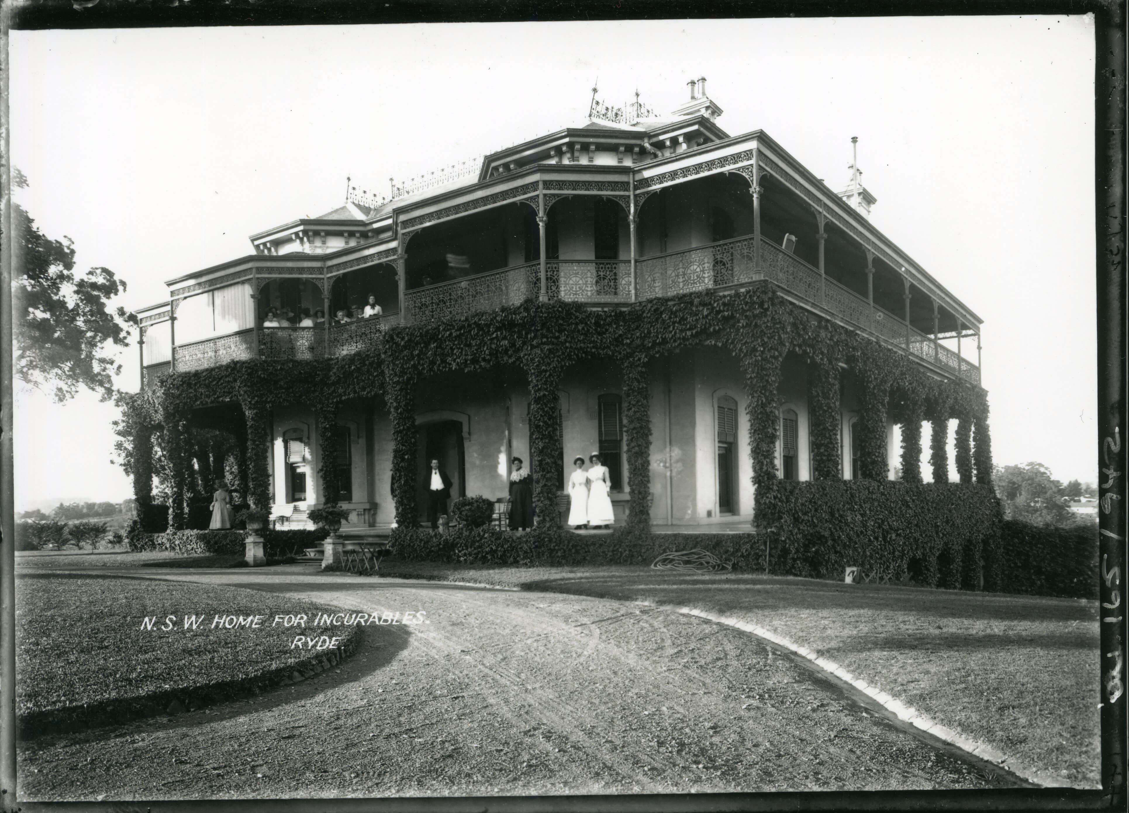 Black and white photo of a large two-story house with a wraparound balcony and veranda. Standing on the veranda and balcony are nurses and patients.