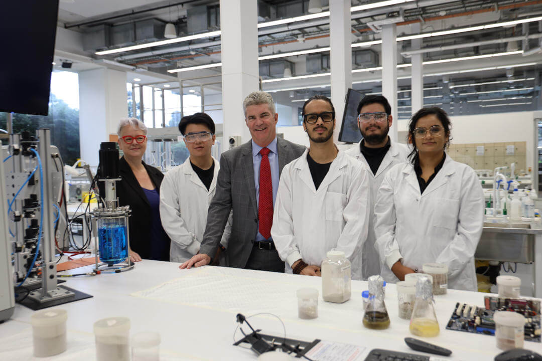 Penny Sharpe and Steve Whan in a lab with 4 students in white lab coats.