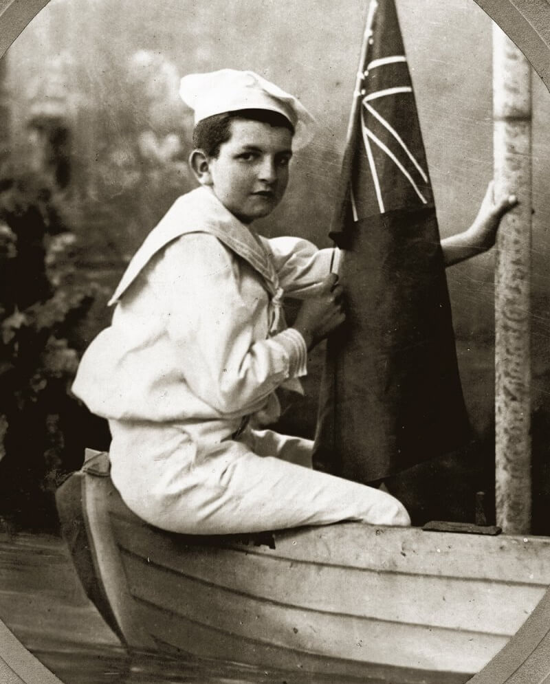 Black and white studio portrait of a young Orry-Kelly, around 8 years old, taken at Cock’s Photographic Studio in Kiama. He is seated on the edge of a small boat marked “Kiama,” wearing a sailor uniform and holding a flag. He looks directly at the camera with a composed expression