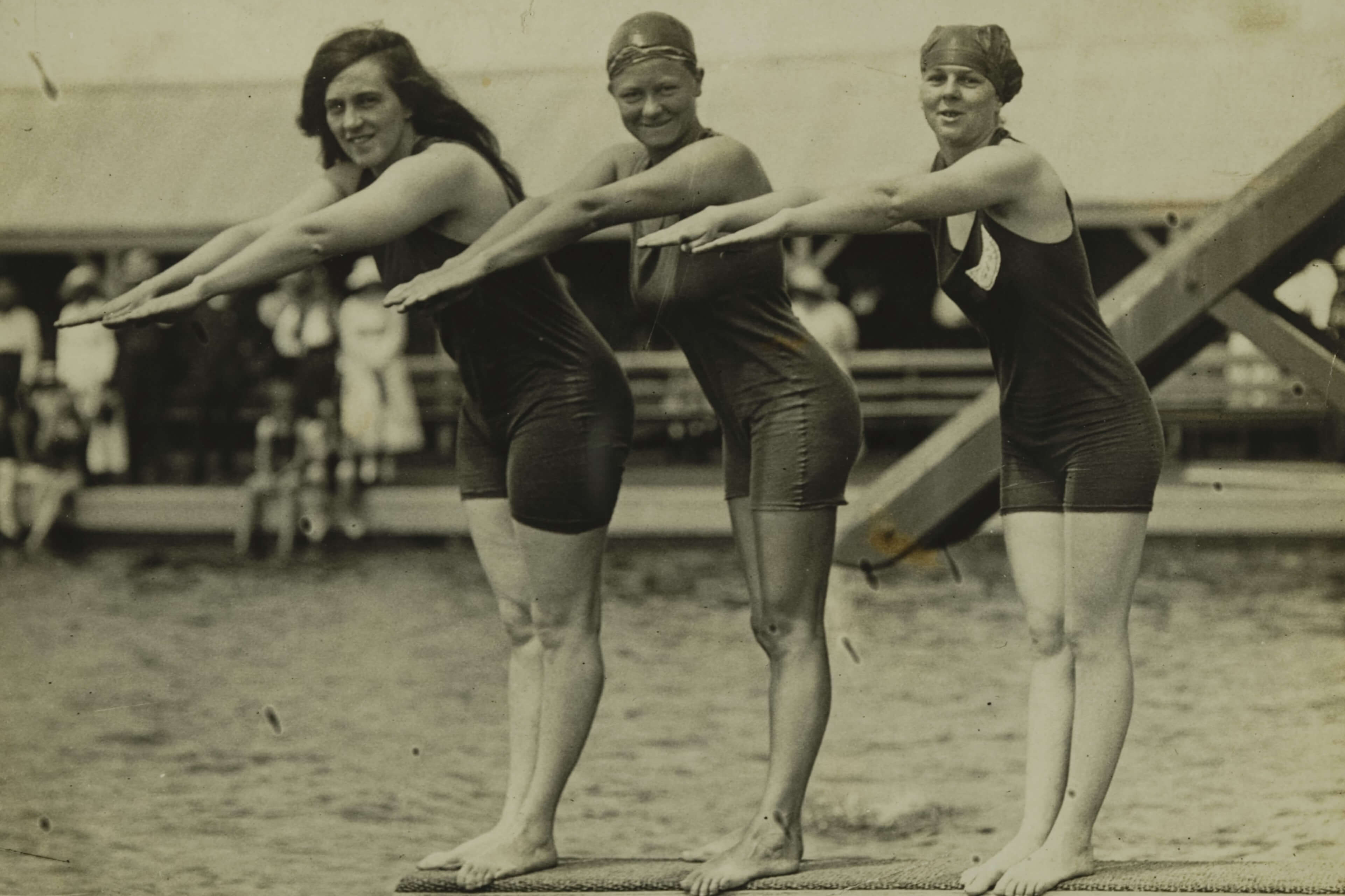 Three female swimmers standing on a diving board in one-piece swimsuits with arms extended forward, preparing to dive. In the background are spectators and a wooden structure near the water's edge.