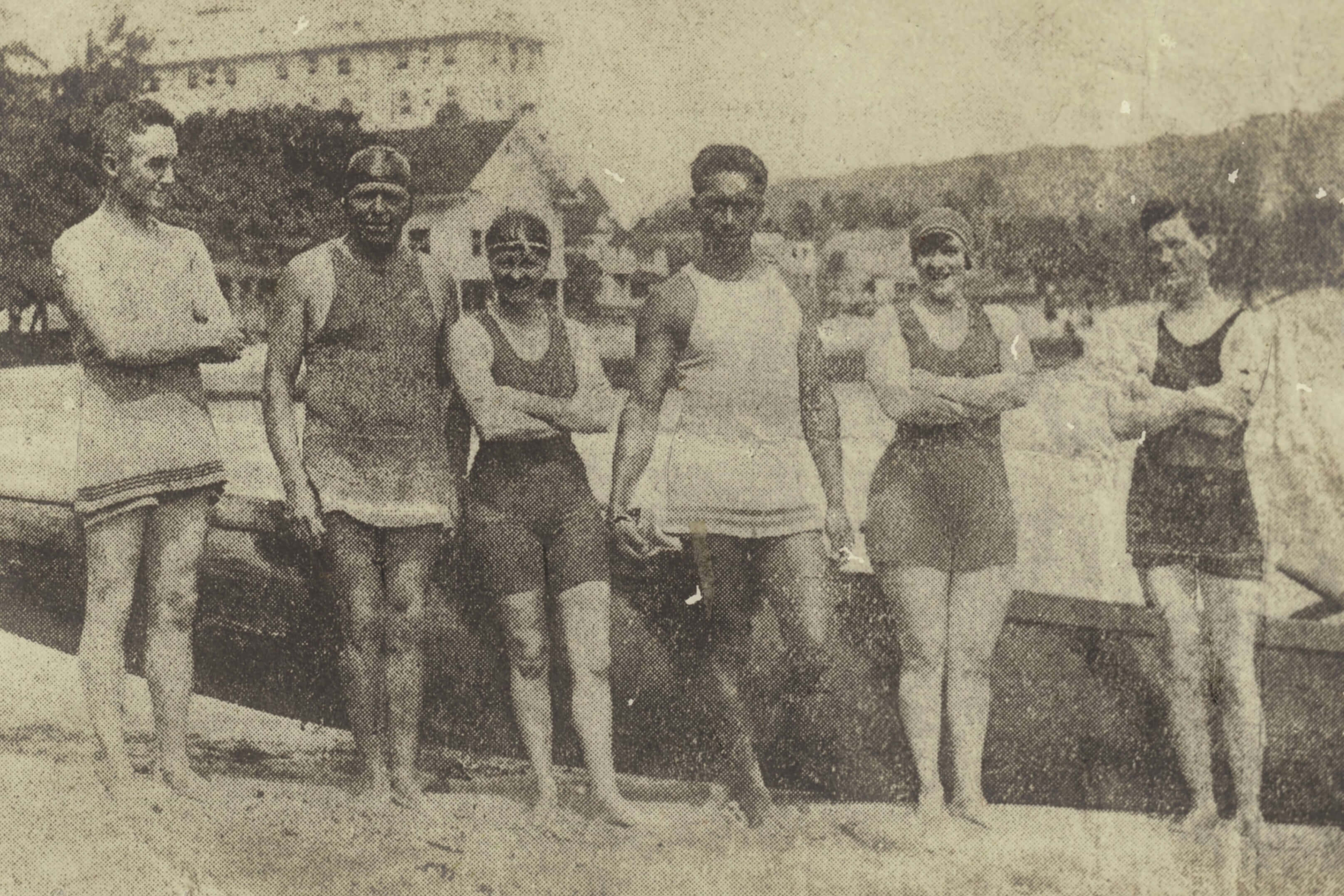 Sepia-toned photograph of 6 individuals, including Mina Wylie, Fanny Durack, and Duke Paoa Kahanamoku, standing on a beach. They are dressed in early 20th-century swimwear and posed in front of an outrigger canoe. A building surrounded by trees and distant hills is visible in the background.