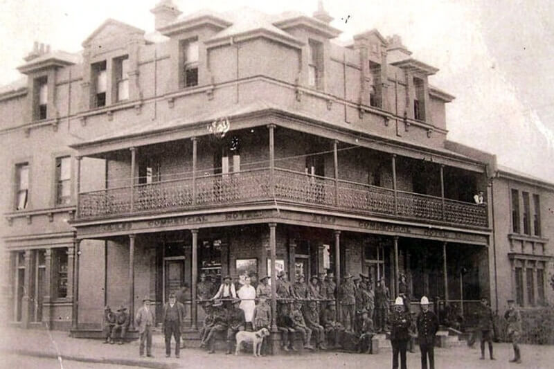 Black and white image of a 3-storey commercial hotel with several people and a dog out the front on the bottom floor. The hotel also has a balcony on the second floor.