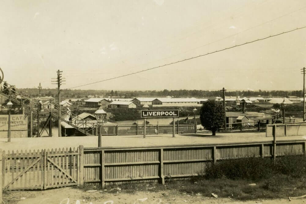 Black and white image of Liverpool army camp. In the middle of the image there is a sign that says ‘LIVERPOOL’. There is a fence in the foreground and a telegraph pole line going across the image.