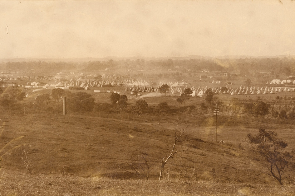 Sepia image of an open grass field. The field is a training camp and in the background are tents and smoke from cooking fires.