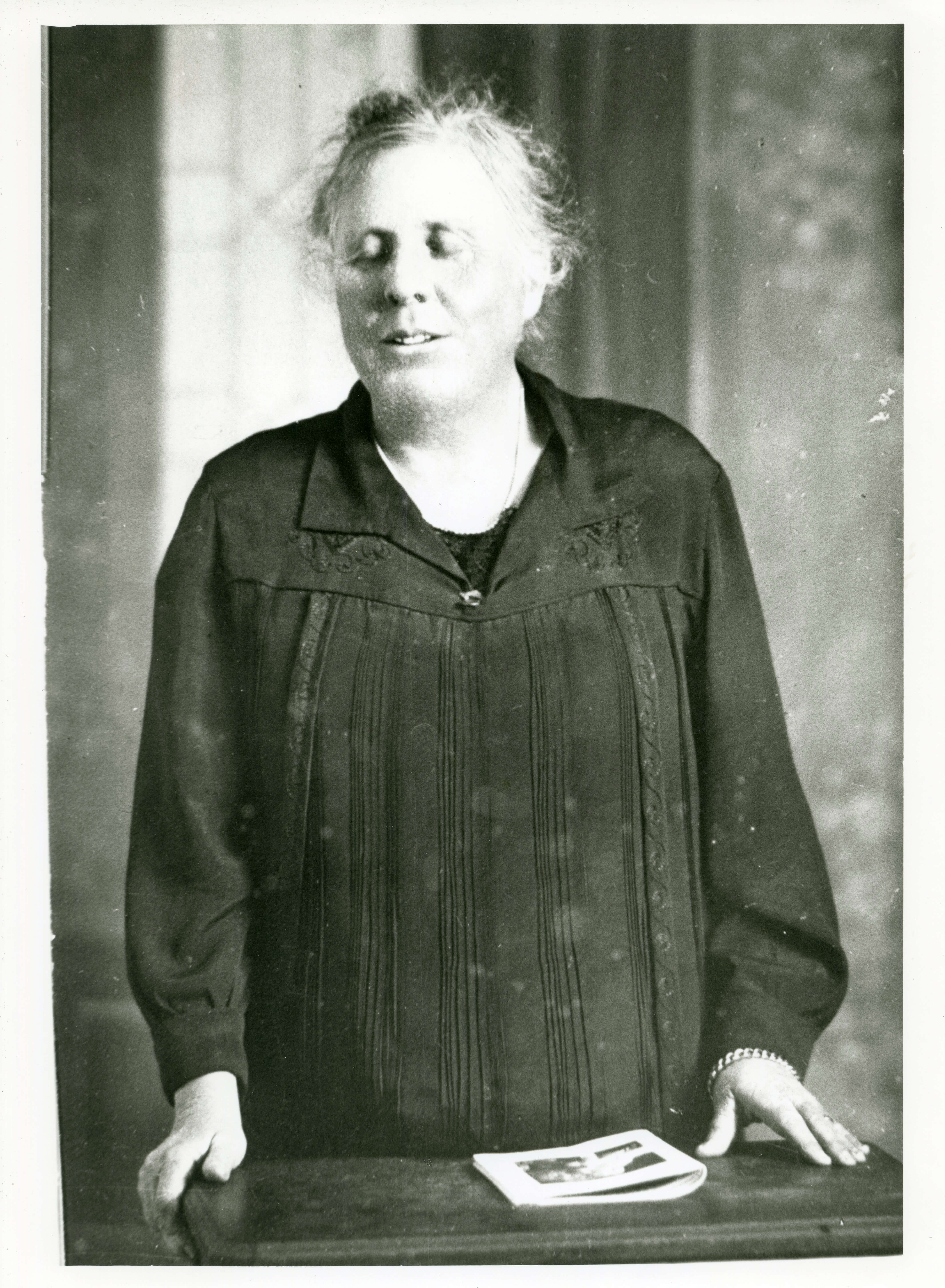 Black and white image of a blind woman. Her hands a resting on a desk beside a piece of paper.