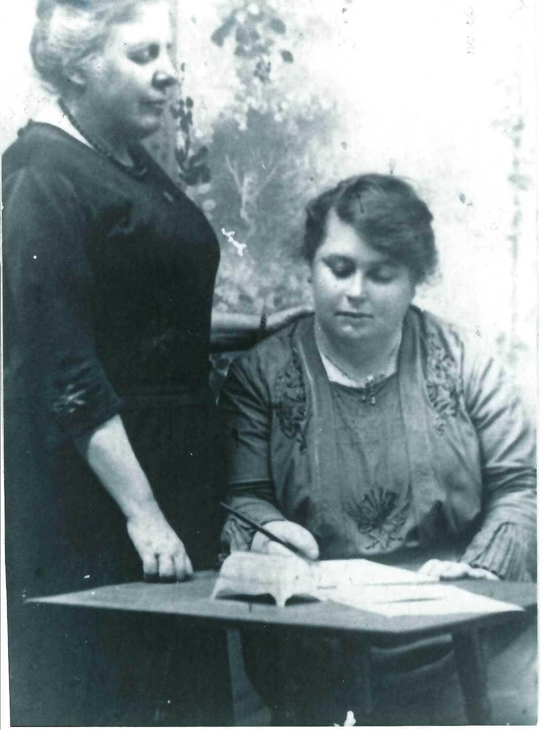 Black and white photograph of Susan, standing with her hand on the shoulder of a female seated pupil.