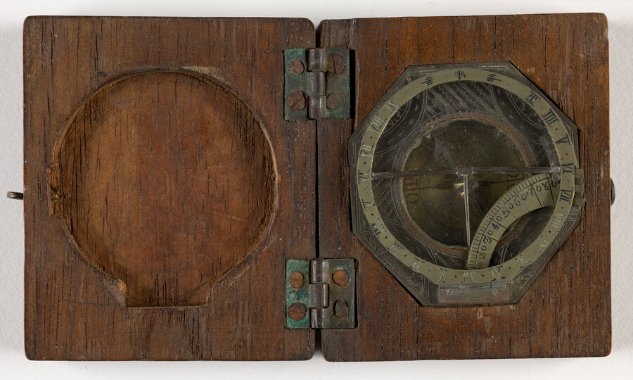 Photo of compass housed in a wooden case with metal hinges. The left side of the case has an indentation for the compass. The right side holds the octagonal compass with a glass cover, marked with degrees and cardinal directions.