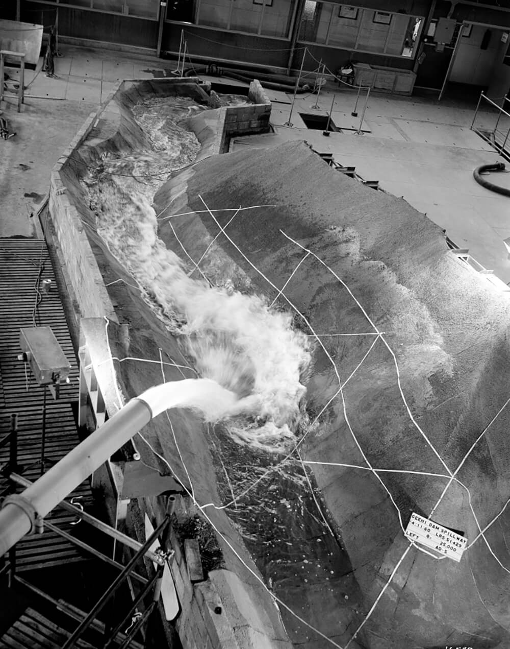 Black and white image of water gushing out of a pipe into a dam spillway model.