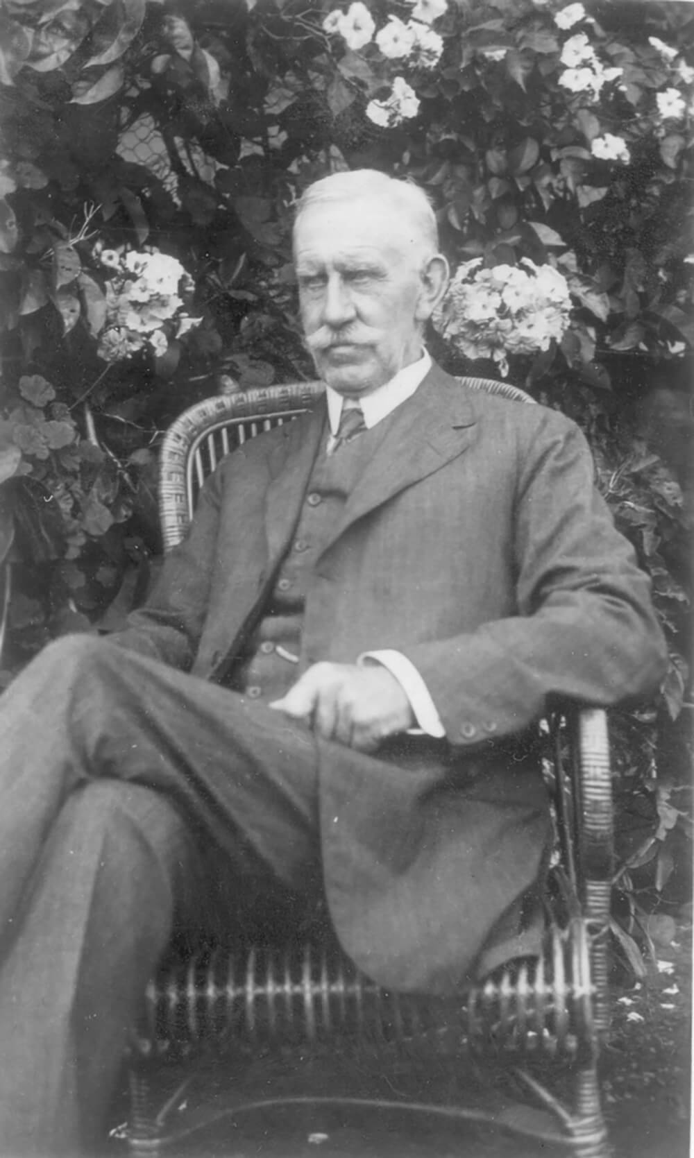 Black and white portrait of a man in a suit sitting on a chair outside with leaves and flowers behind him.