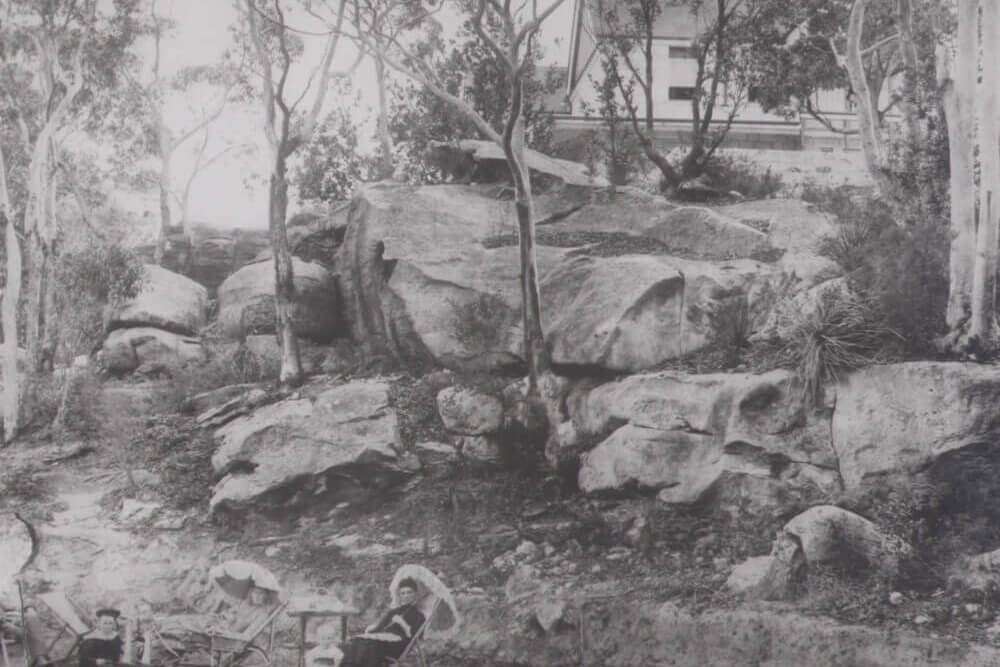 Black and white image of a family picnicking in the foreground with trees and large rocks behind them. On top of the rocks is a house.