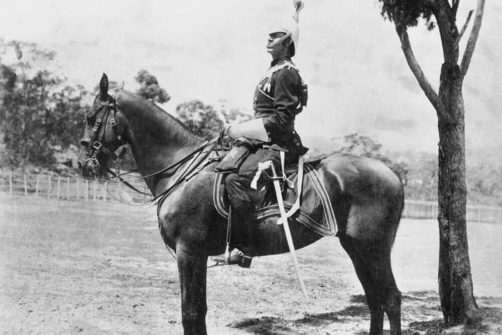 Black and white image of a man sitting on a horse. The man is wearing an army uniform and has a sword hanging off his left leg. There is a tree next to them on the right and forestry in the background.