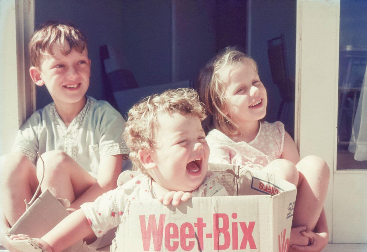 Three children sitting on a porch, with one child inside a large Weet-Bix cereal box. The other two children are seated directly on the porch. The image is taken outdoors in natural sunlight.
