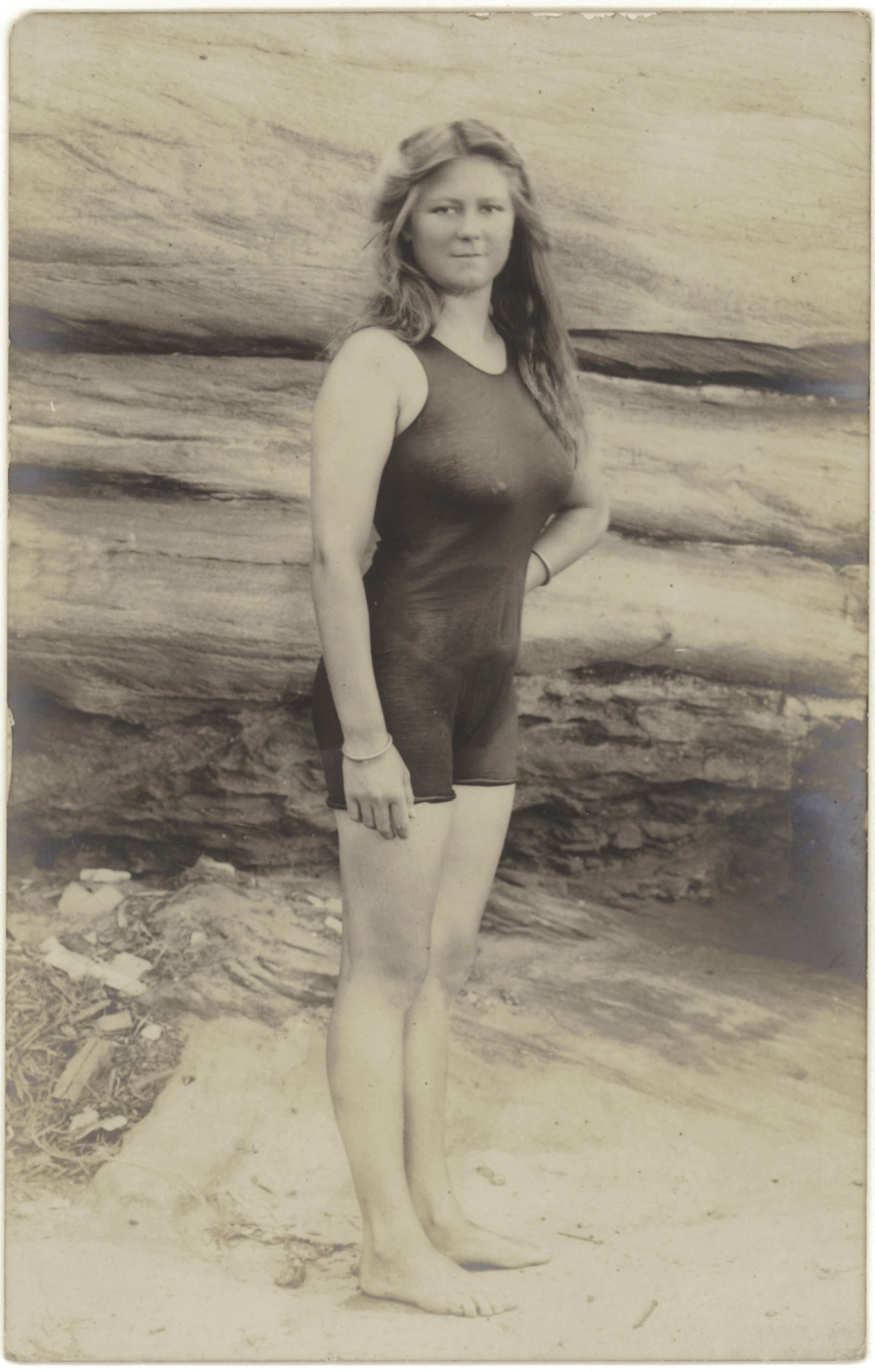 Sepia-toned photograph from 1910 showing Wilhelmina (Mina) Wylie standing barefoot on a beach, wearing a dark, sleeveless one-piece swimsuit. Her long hair is loose, and she poses with one hand on her hip in front of a rocky background.