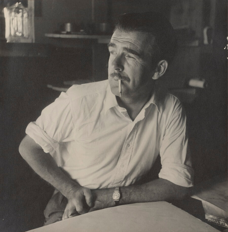 A black and white image of a man with his arm resting on a desk and a cigarette in his mouth.