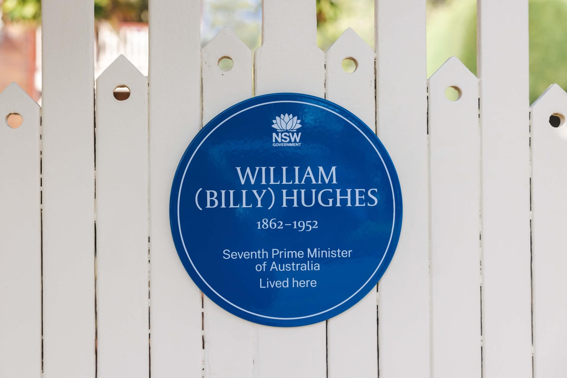 A blue plaque for Billy Hughes on a white picket fence gate.