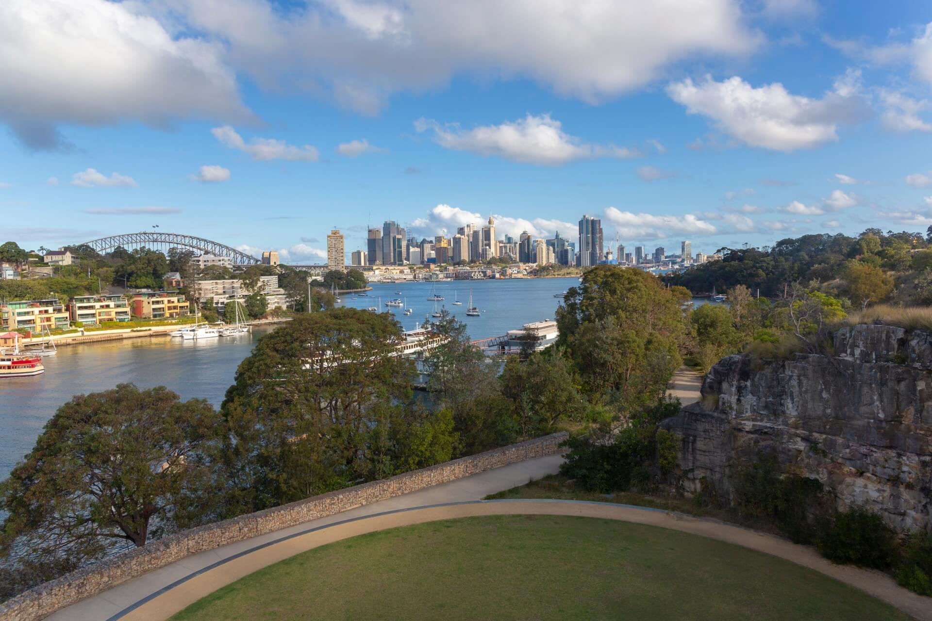 View of a riverside park with a blue harbour, houses, boats and the city skyline in the background