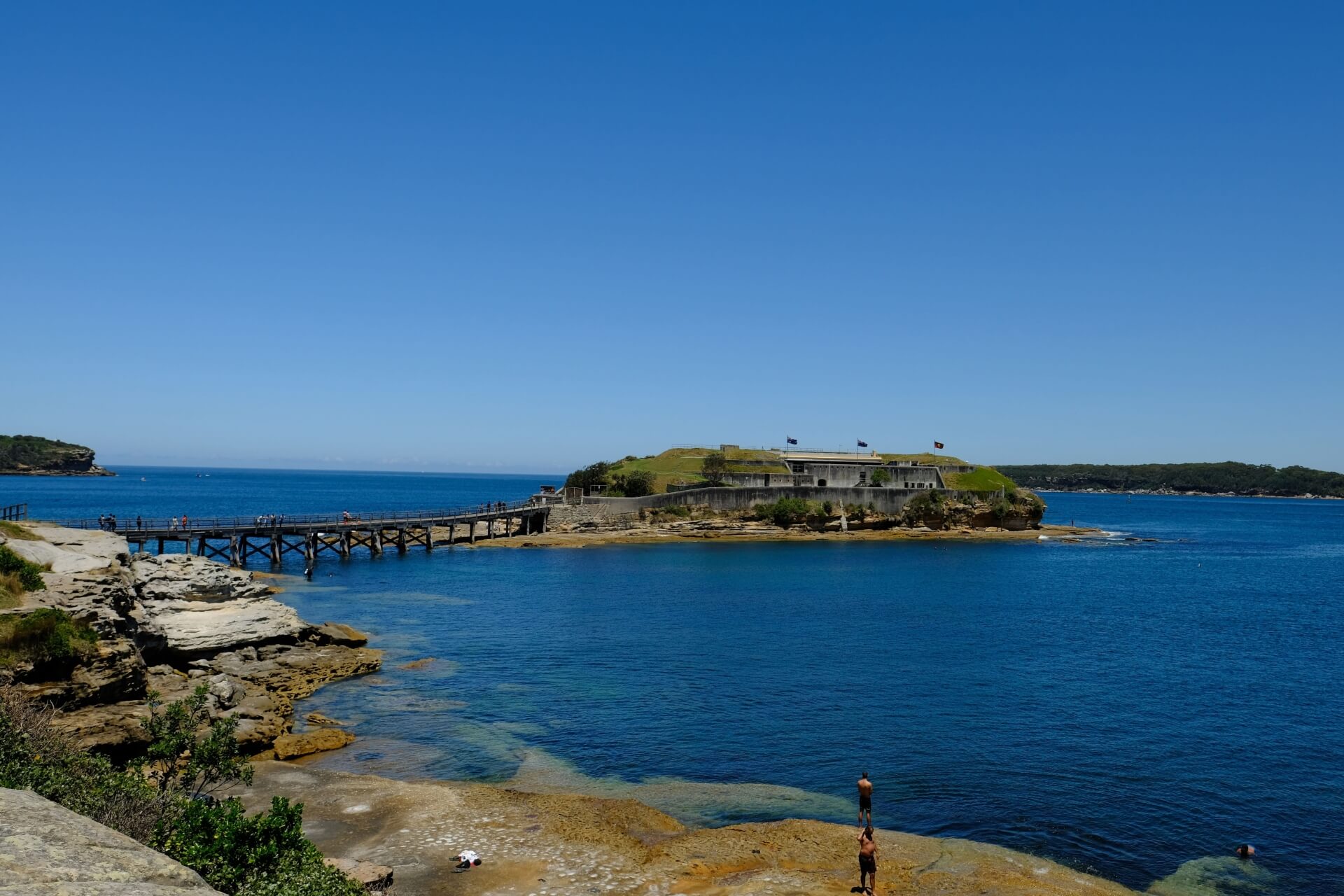 A small island with buildings on it, accessible by a timber footbridge, surrounded by blue ocean