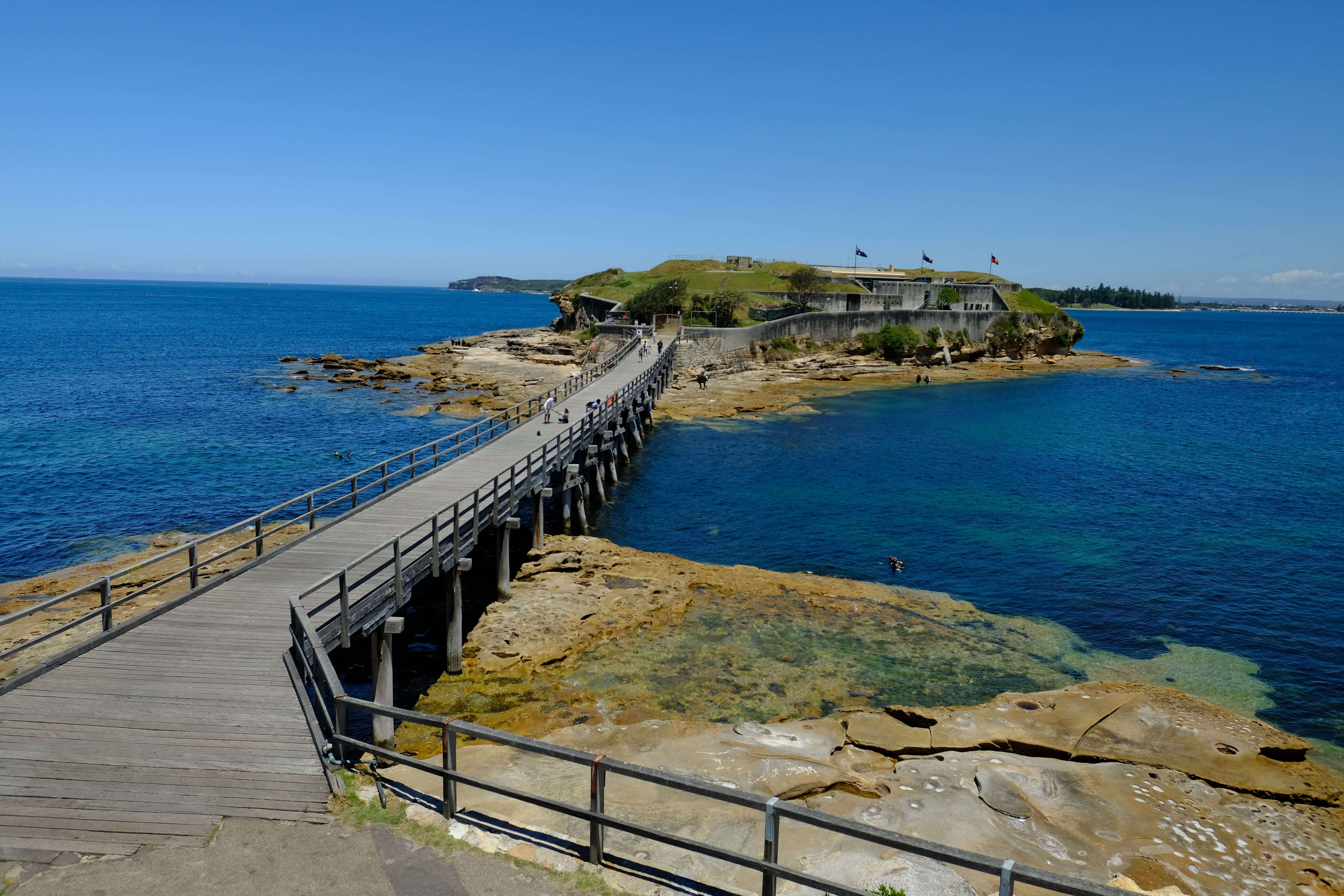 A wide timber pedestrian bridge extending over blue water to a small island surrounded by the sea