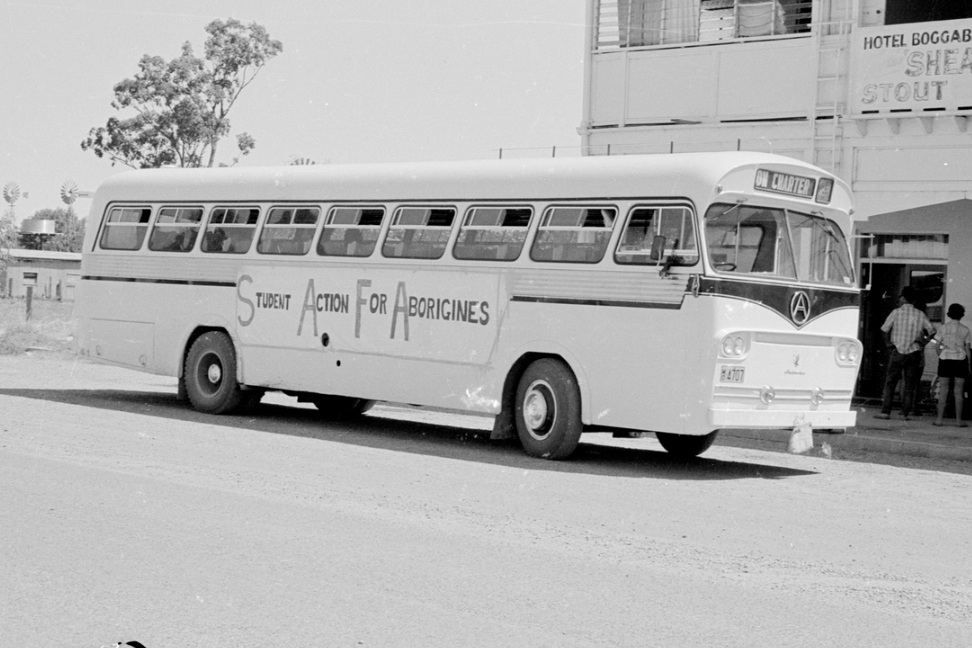 Black and white photo of a large bus bearing a hand-made sign 'Student action for Aborigines'. The bus is parked outside the Hotel Boggabilla and there is a small group of people standing at the hotel entrance.