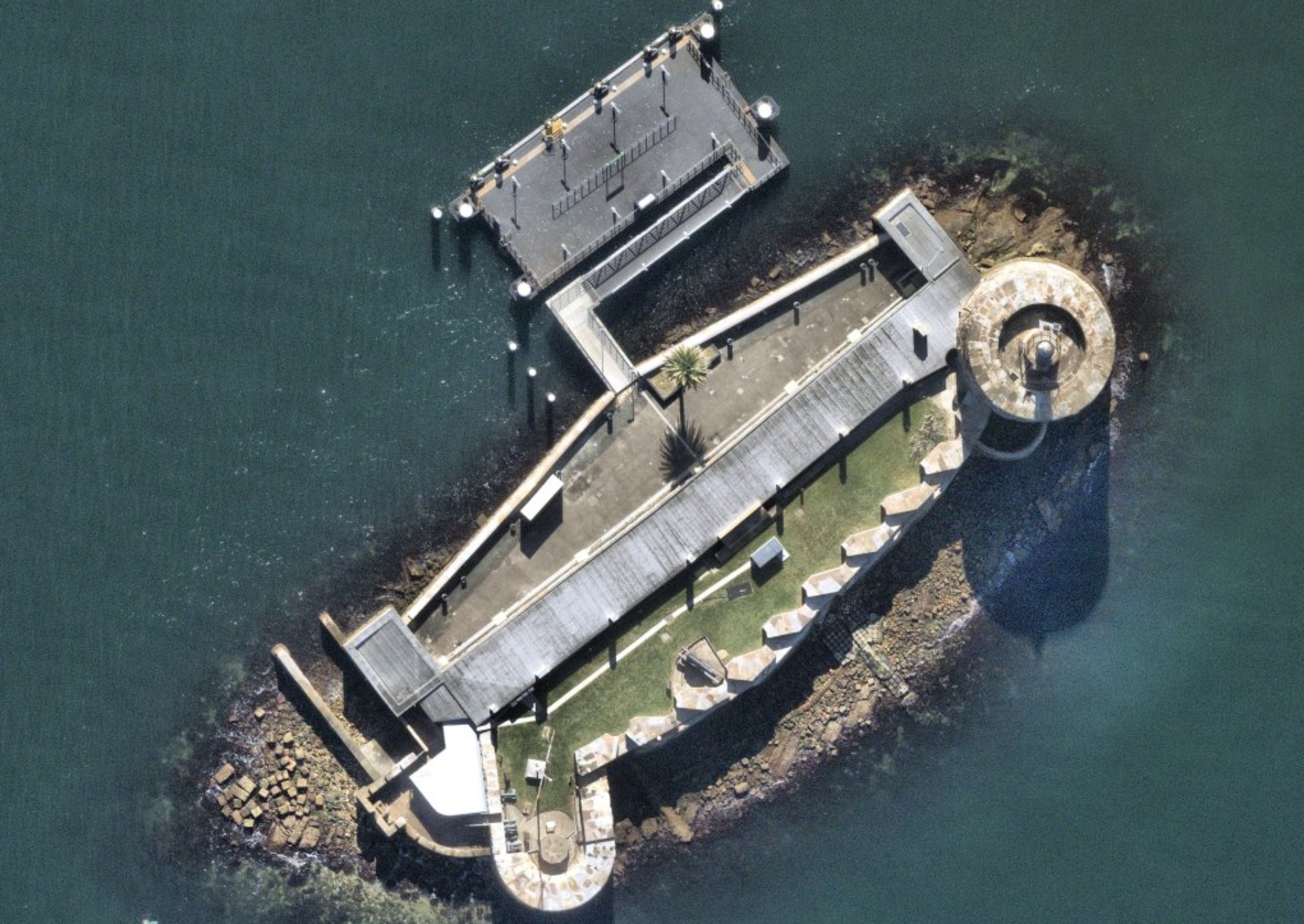 Aerial view of a small oval-shaped island with a rocky shore, wharf, buildings and landscaping