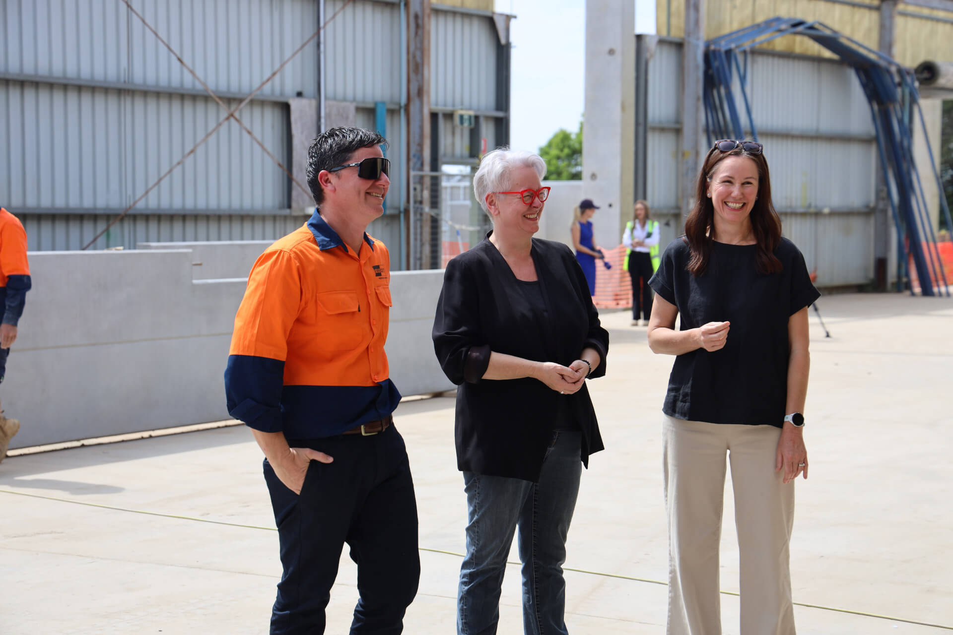 Three people, one in a bright safety shirt, stand in a large warehouse-type space.