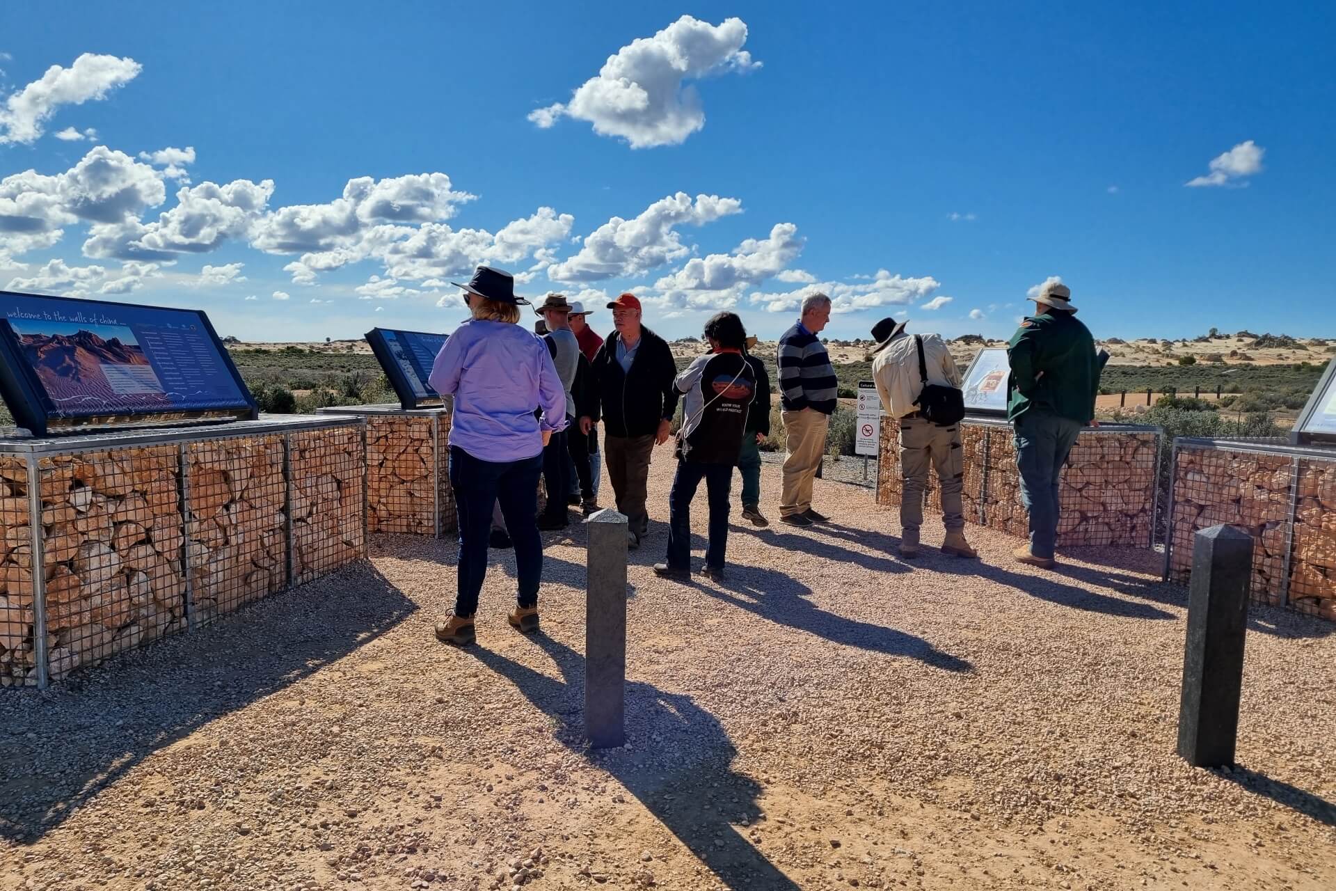 A group of 9 people walk on a gravel path inspecting national park signs on a sunny day