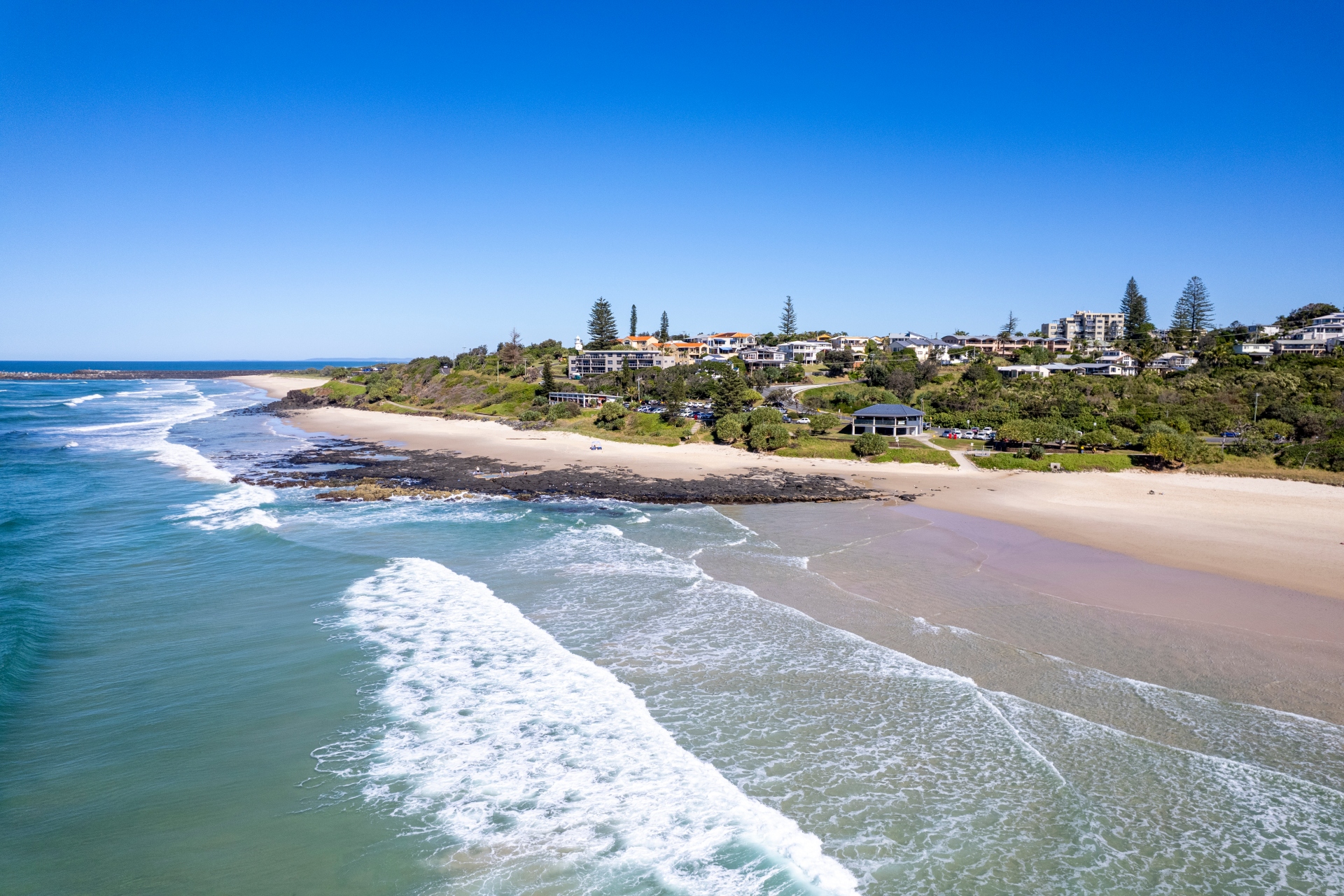 Trees and houses flank a wide, golden sandy beach next to blue-green ocean and waves breaking