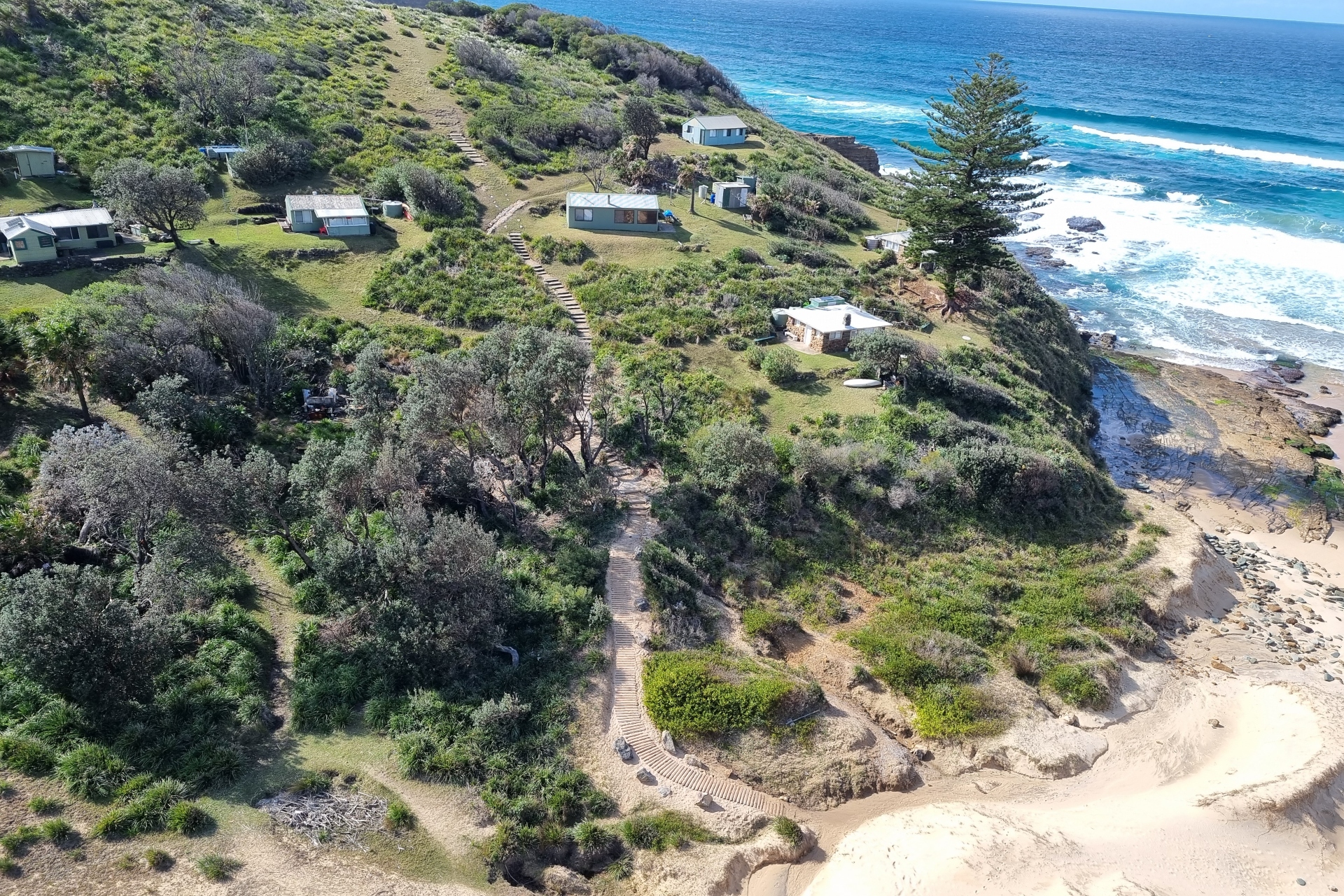 A winding staircase leading from a headland dotted with trees and houses down to a golden sandy beach next to blue ocean