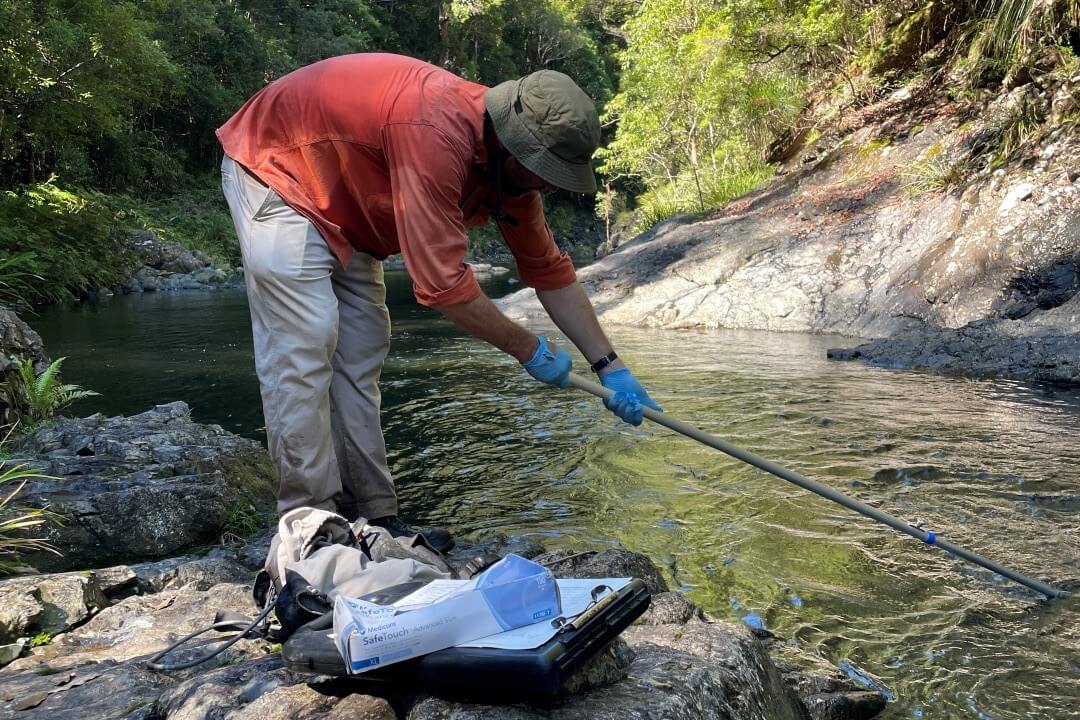 Person in red shirt, light coloured pants and hat bending over stream with equipment in water and sample jars in foreground.