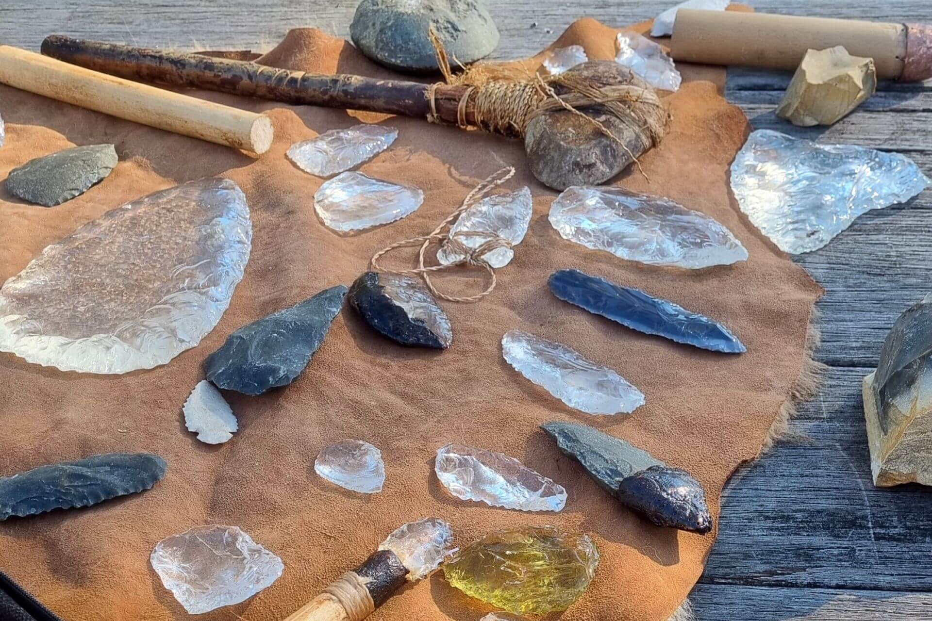 Collection of Aboriginal cultural objects displayed on a leather hide, including stone and glass spear points, blades, scrapers and cutting tools. Wooden-handled implements, stone artefacts and flakes are arranged on a timber surface, demonstrating traditional tool-making techniques and the adaptation of both natural and recycled materials.