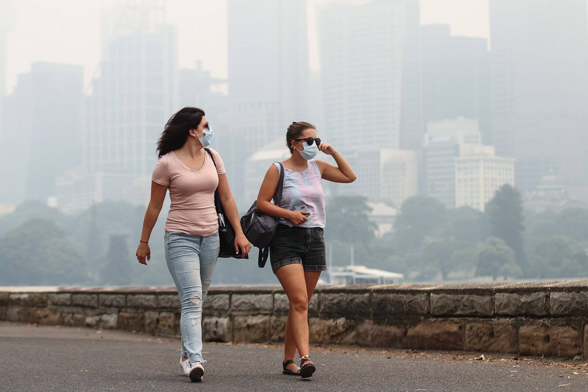 Two people walking along asphalt path by Sydney Harbour, wearing masks, with city lost in smoke haze behind them. 