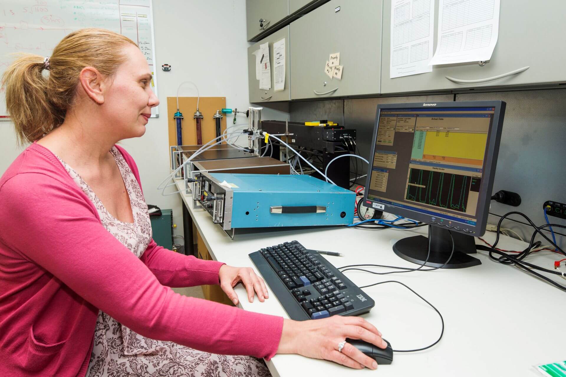 A person sits at a laboratory workstation adjusting a blue analytical instrument connected by tubing to calibration equipment. A computer monitor displays graphs and measurement data, while laboratory cabinets and additional instruments are visible in the background.
