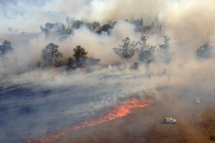 Aerial view of a wildfire with visible flames and thick smoke, spreading across a landscape with trees and a road, where two vehicles are present near the fire line.
