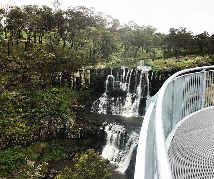 Scenic view of a multi-tiered waterfall cascading down rocky cliffs, surrounded by lush green vegetation, as seen from a metal observation platform at Lookout 3 in Guy Fawkes River National Park.