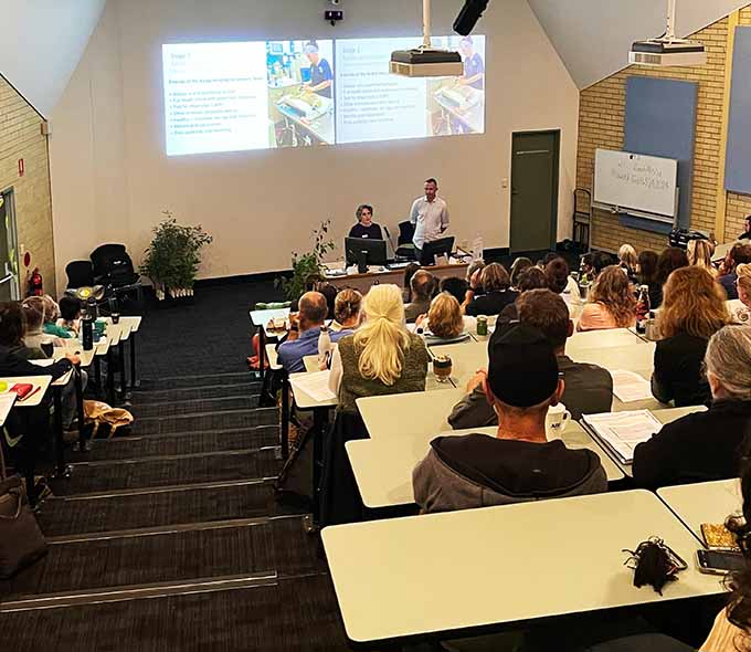 A conference room scene with attendees watching a presentation delivered by two speakers