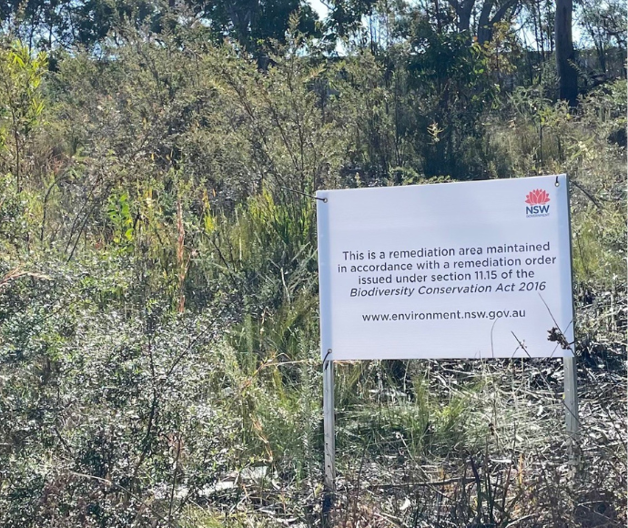 White sign posted in a bushy, natural area. The sign is placed in the foreground, surrounded by dense vegetation typical of Australian bushland, including various shrubs and small trees.