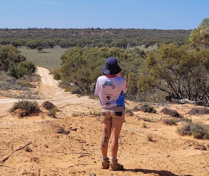 Ranger in bucket hat and shorts, back to camera, staring down dusty road