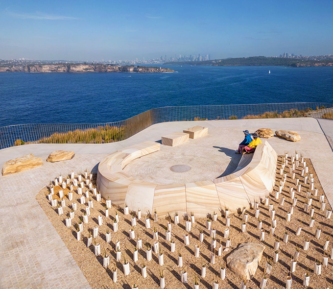 A person is seated on a curved bench by the sea, surrounded by vertical wooden stakes