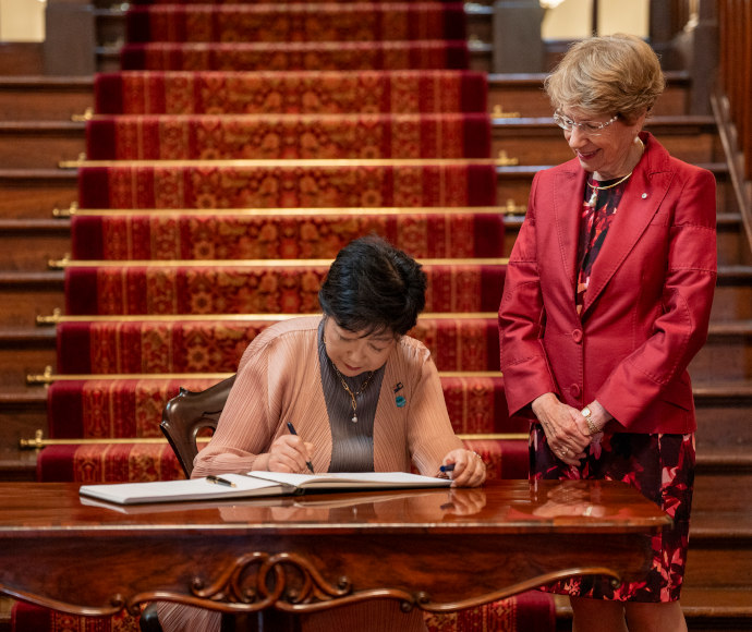 A well-dressed woman sitting at an ornate desk signs a document