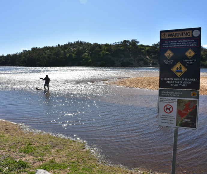 Warning sign at lakeside beach; person wading in water; sun glinting off lake surface; forested hills beyond; safety hazards indicated.