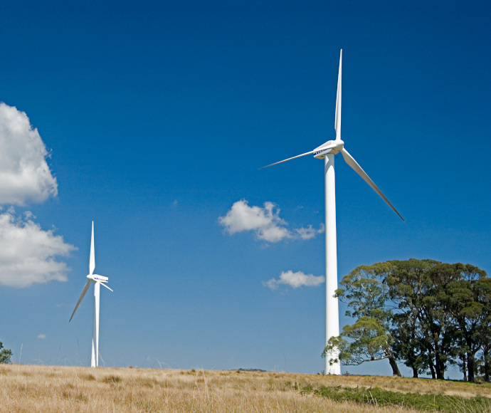 Two wind turbines on top of a dry hill. There are some trees in the foreground and a blue sky with fluffy white clouds behind the turbines.